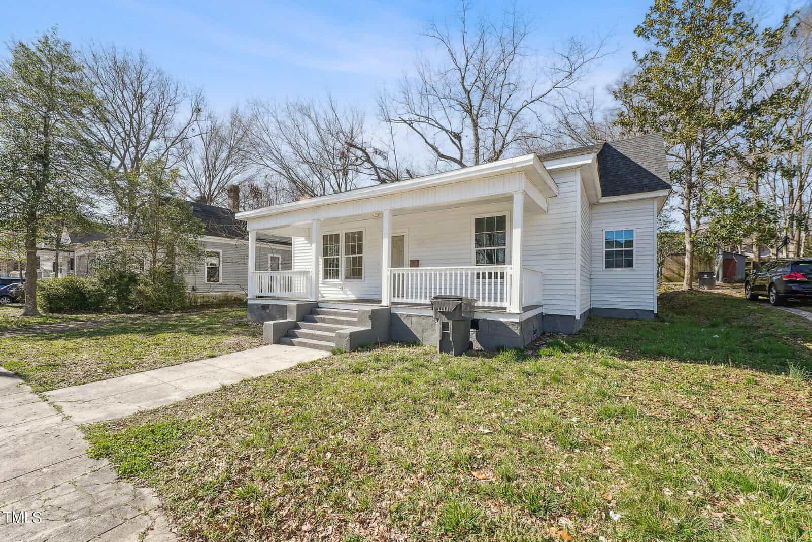 A small white house with a front porch and three steps leading up to it, located on a sunny day with clear skies and surrounded by bare trees and a neatly maintained lawn.