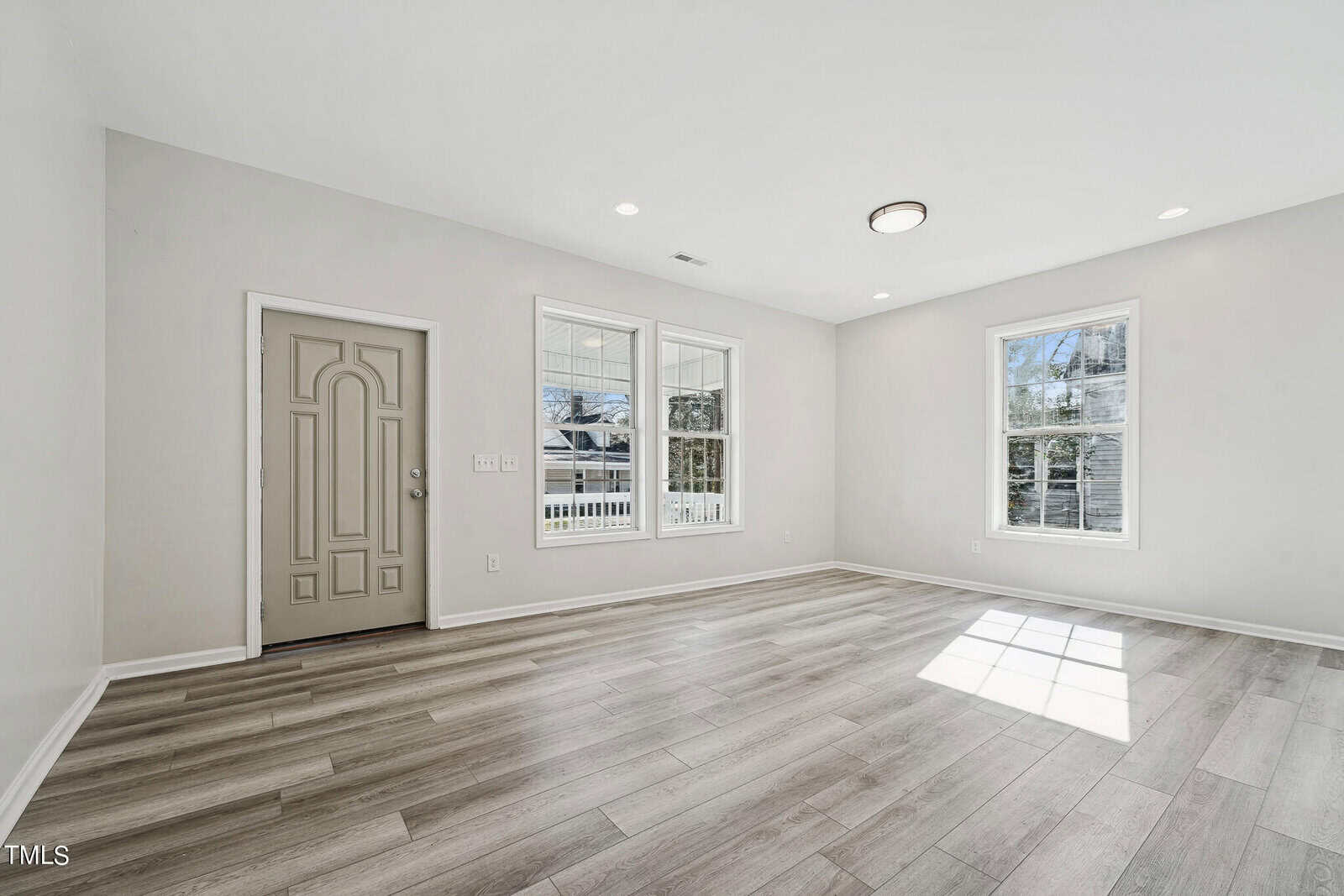 A bright, empty room with a wooden floor, white walls, recessed lighting, and a front door at one end. there are windows with a view of trees and a glass door leading outside.