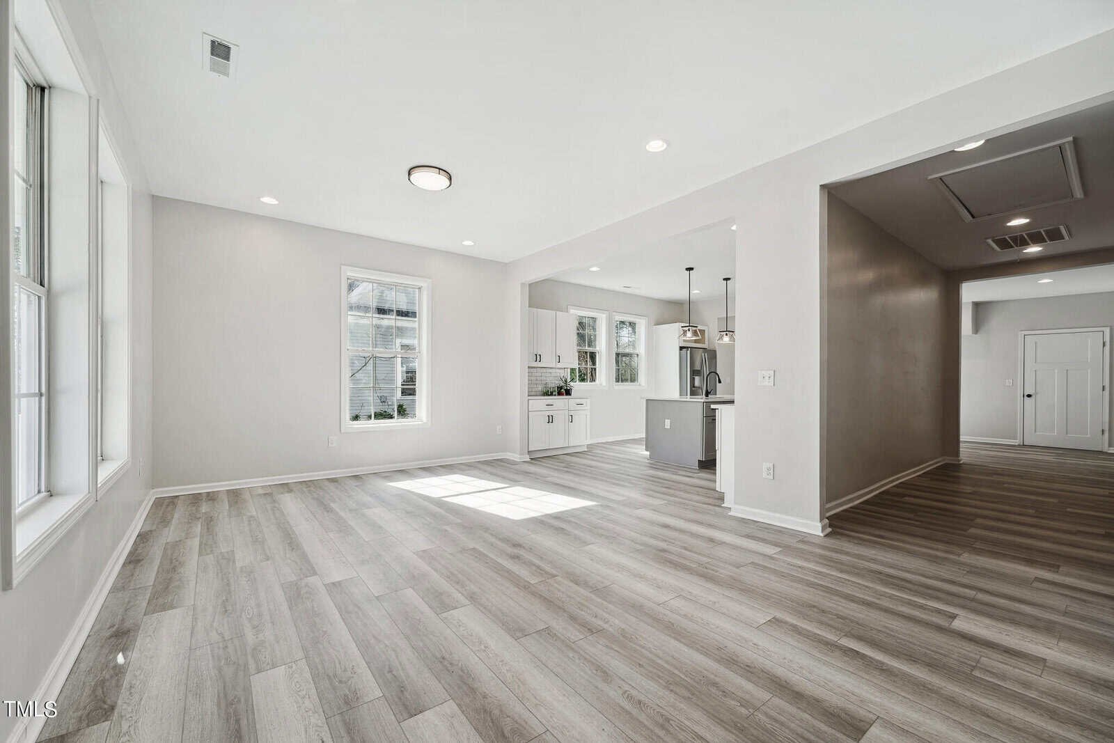 Spacious, modern living room with light hardwood flooring, large windows, and an open floor plan leading to a white kitchen with a bar counter. neutral walls and ample natural light.