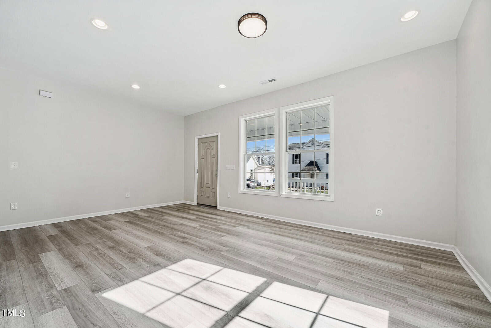 Empty modern living room with gray wood floors, white walls, a front door, and large windows letting in natural light.