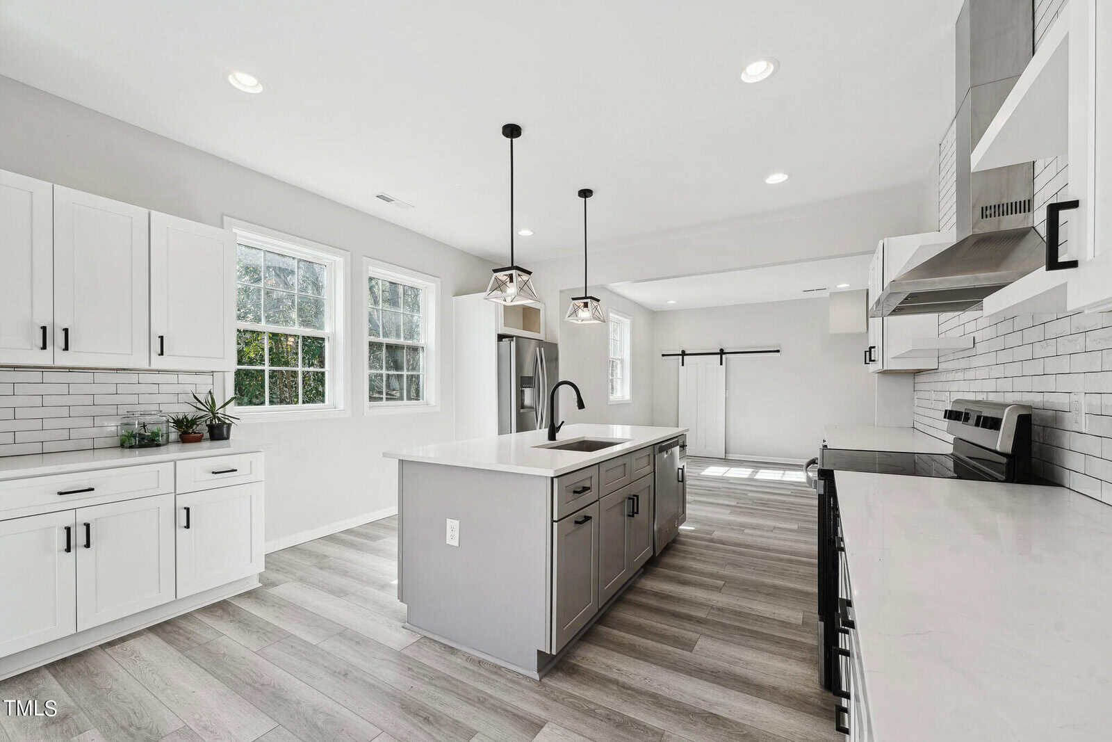 Modern kitchen with white cabinets, subway tile backsplash, a central island with a sink, and hanging black pendant lights. windows provide natural light, complementing the light hardwood floors.