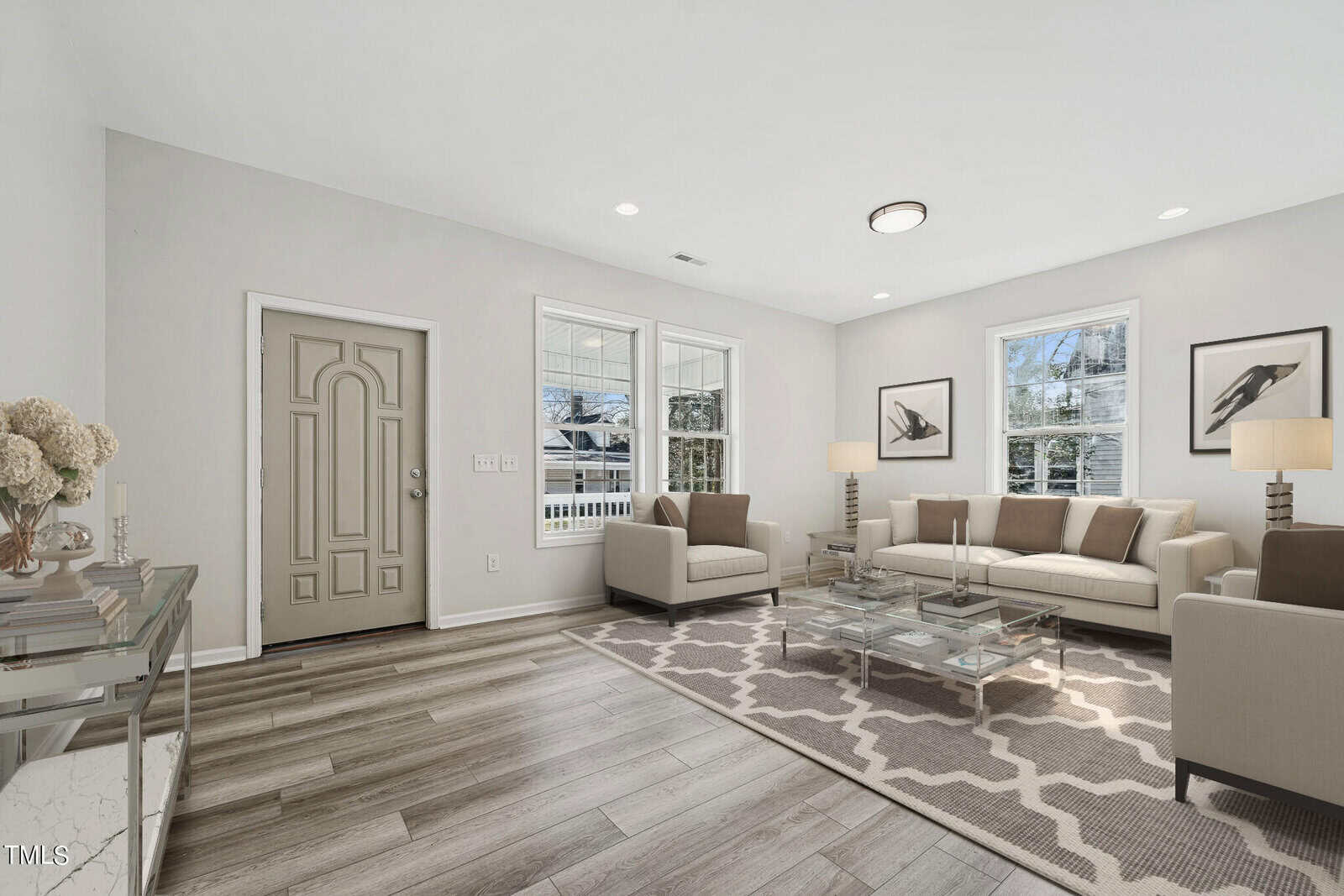 A modern living room with light hardwood floors featuring two beige sofas, glass coffee tables, a geometric area rug, and a view of snowy trees through sliding glass doors.
