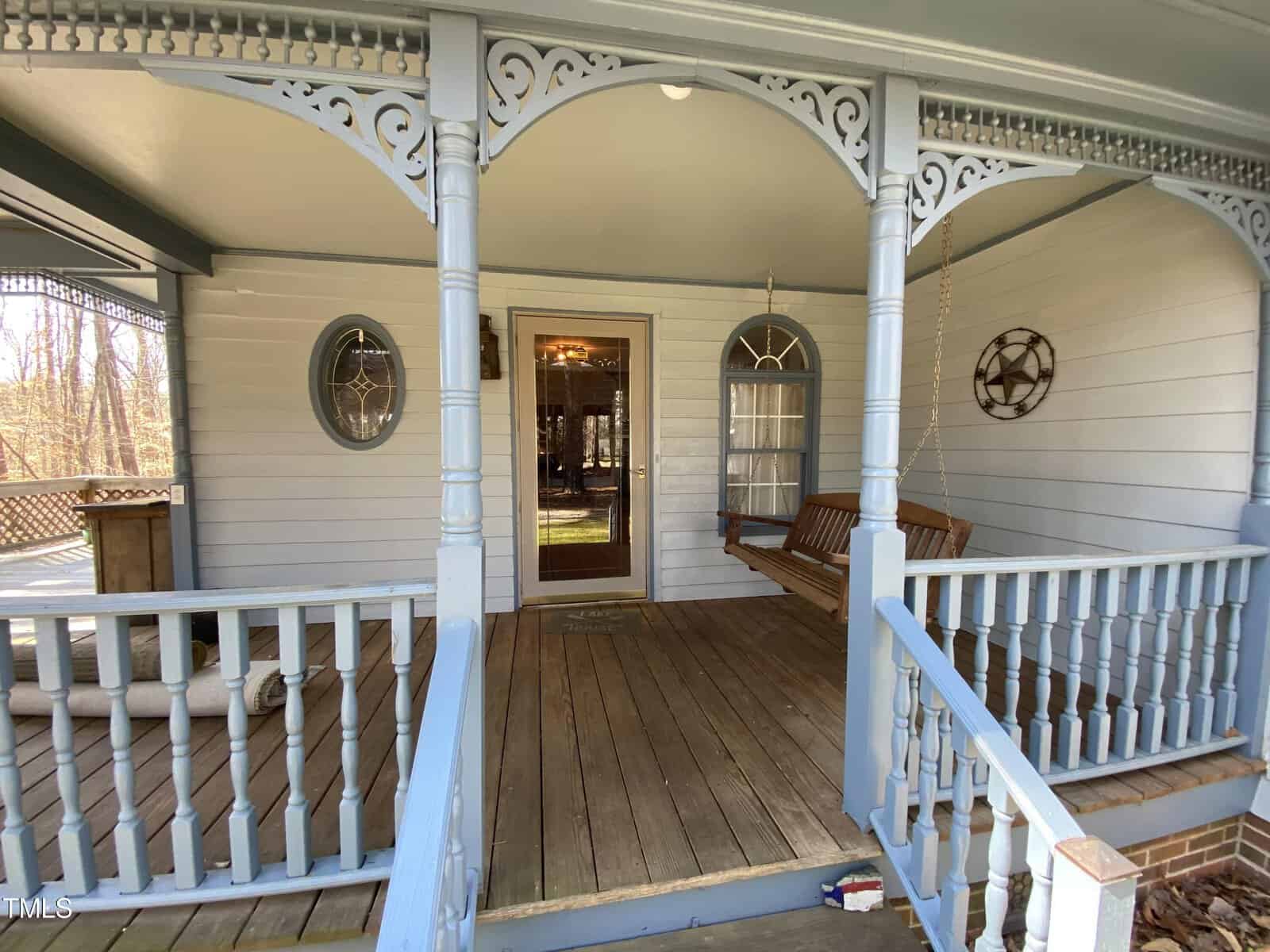 A wooden porch with decorative white railings and archways. there is a wooden bench to the left and two circular wall decorations. the glass door to the house is open, showing an interior view.