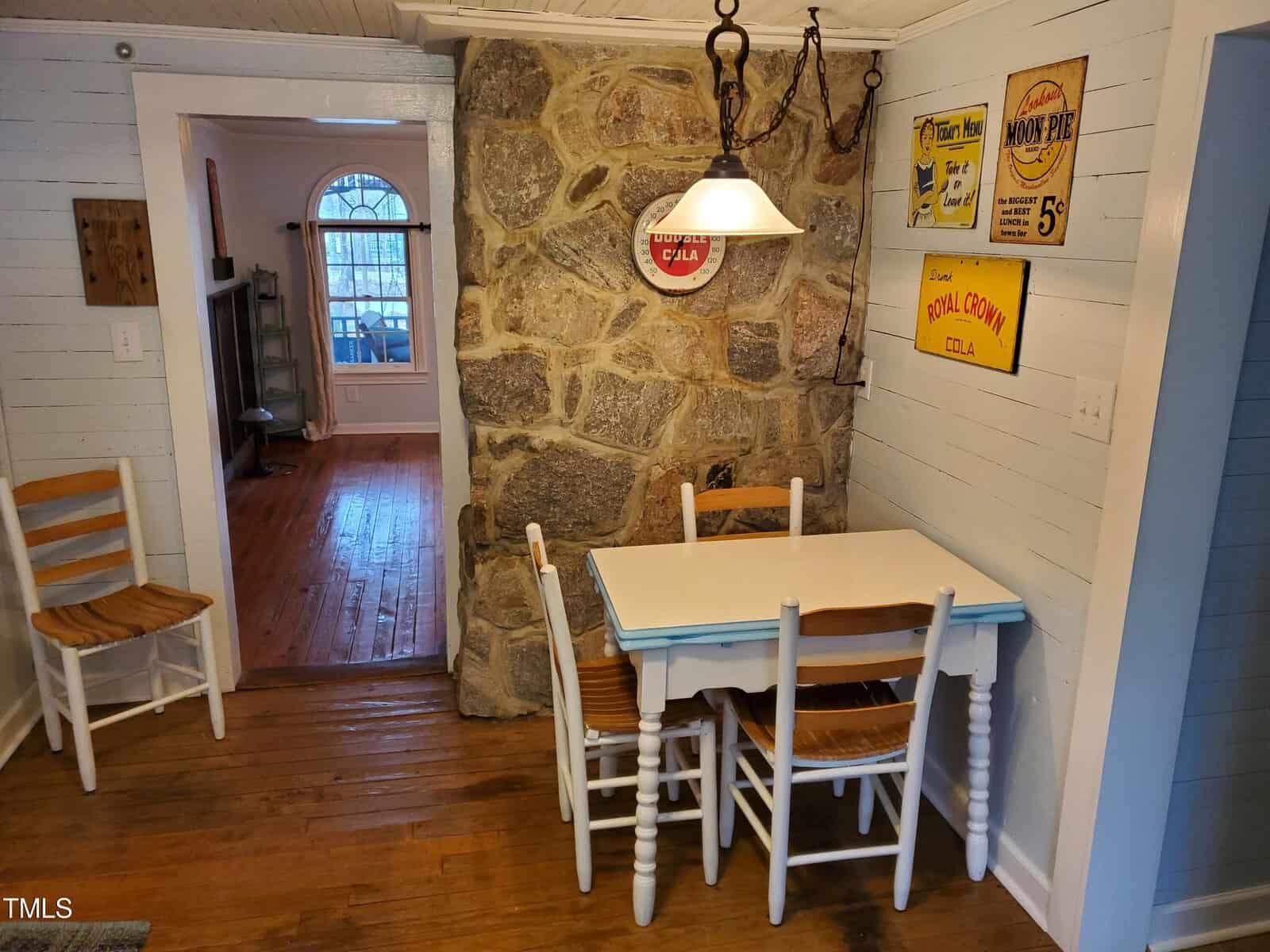 A cozy dining area featuring a small white table with chairs, set against a rustic stone wall adorned with vintage signs. a glass door on the left provides natural light.