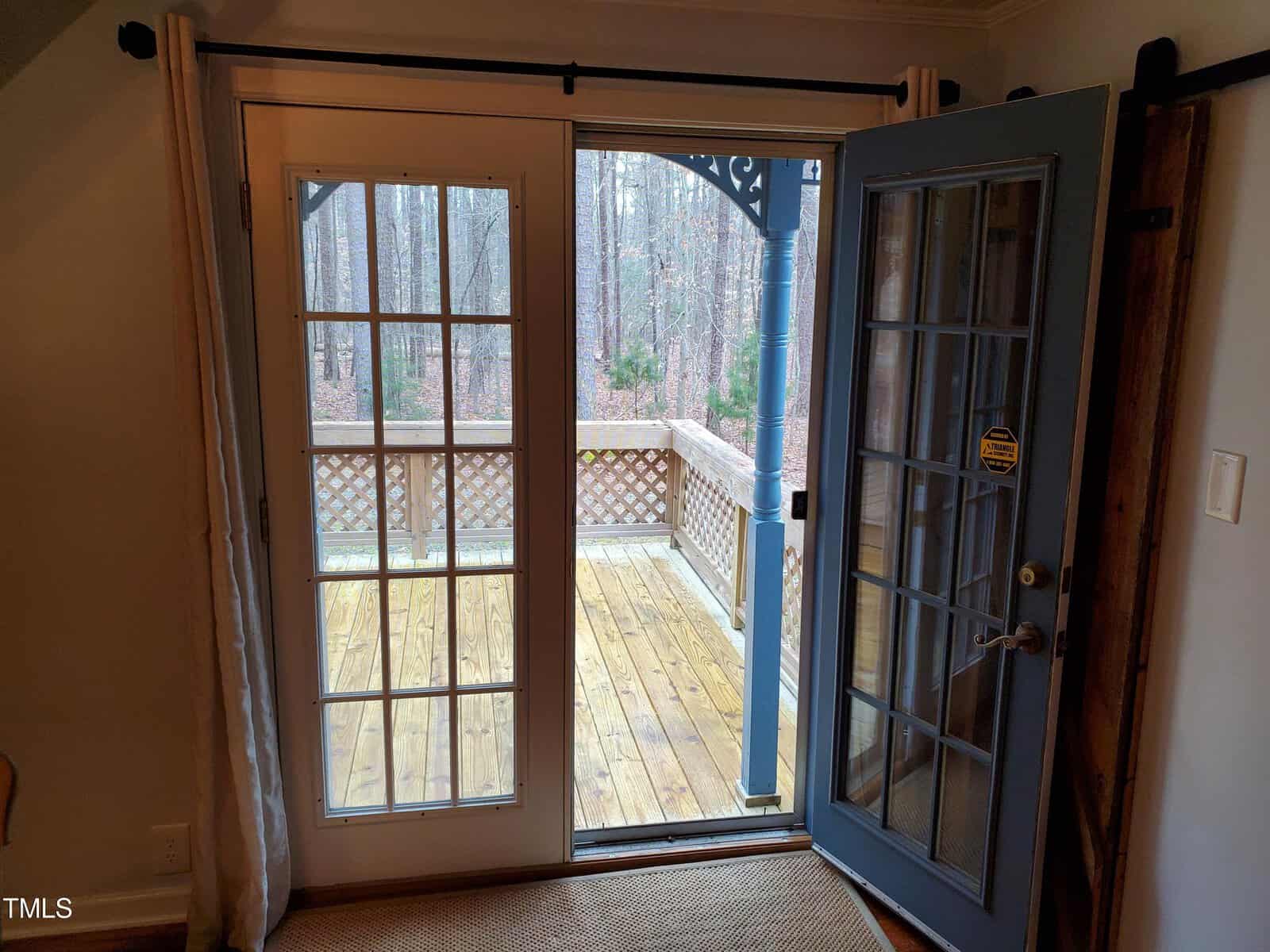 A view from inside a room looking out through an open french door onto a wooden deck with a railing, surrounded by a forest of bare trees. light curtains frame the door on either side.