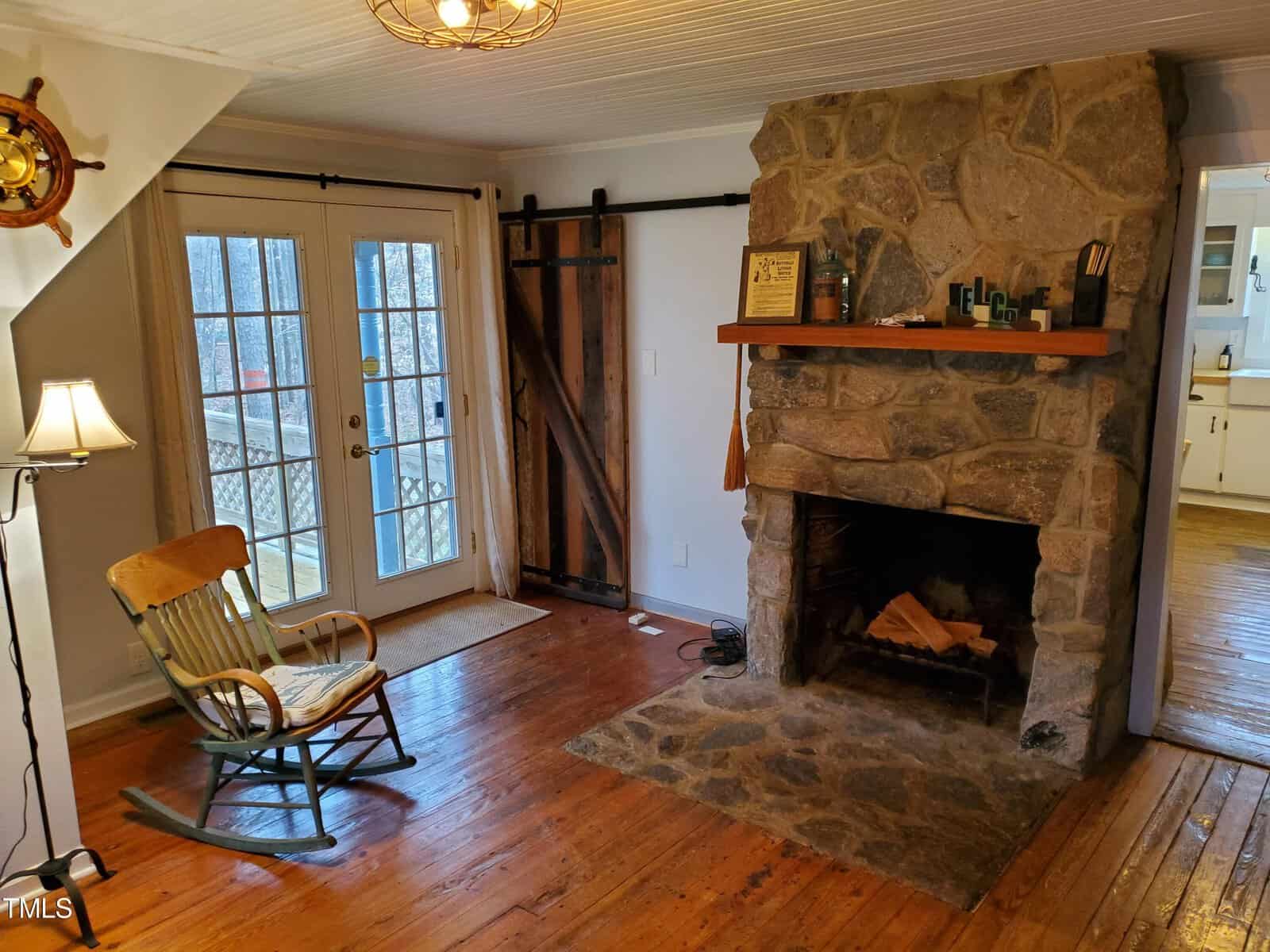 A cozy room featuring a stone fireplace with a wooden mantel, a rocking chair, and a sliding barn door next to glass-paned doors leading outside. hardwood floors and a ceiling light complete the space.