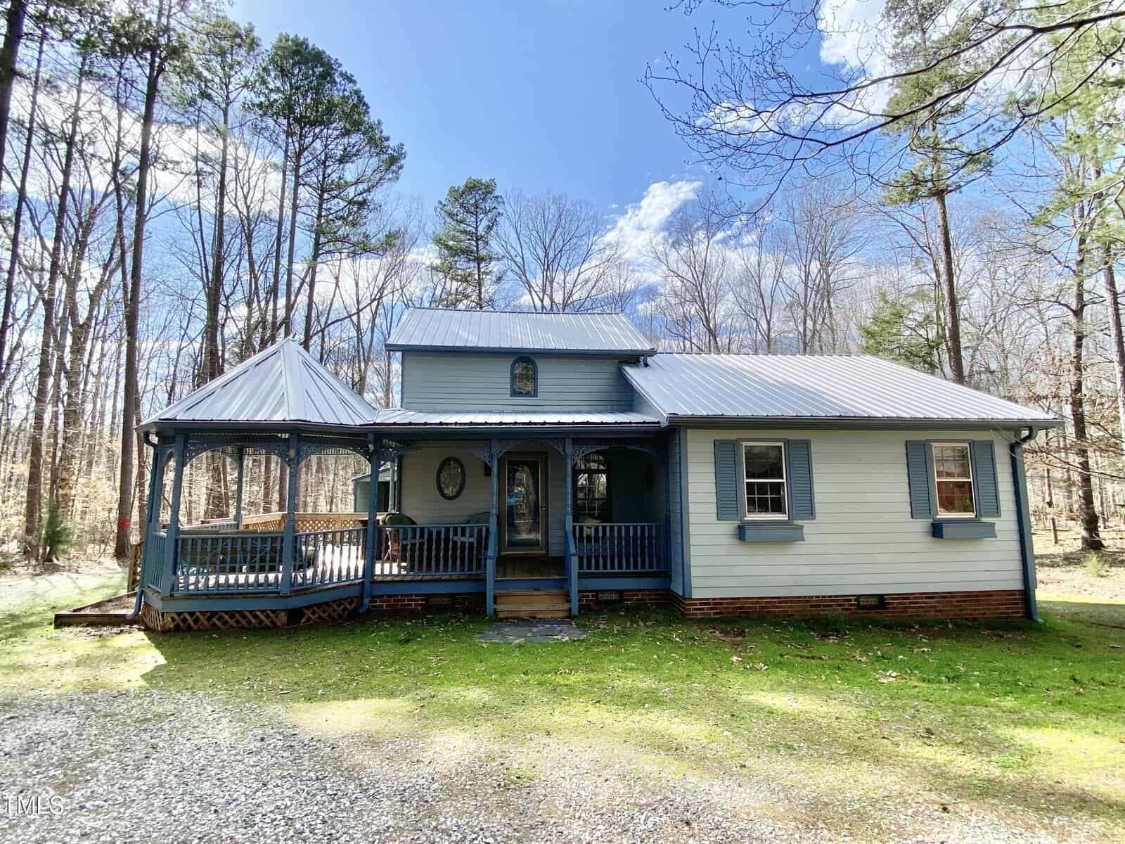 A quaint blue house with a gray roof, located at 358 Pool Rock Shores Lane in Henderson, NC, featuring a wrap-around porch and a screened gazebo, nestled in a wooded area with