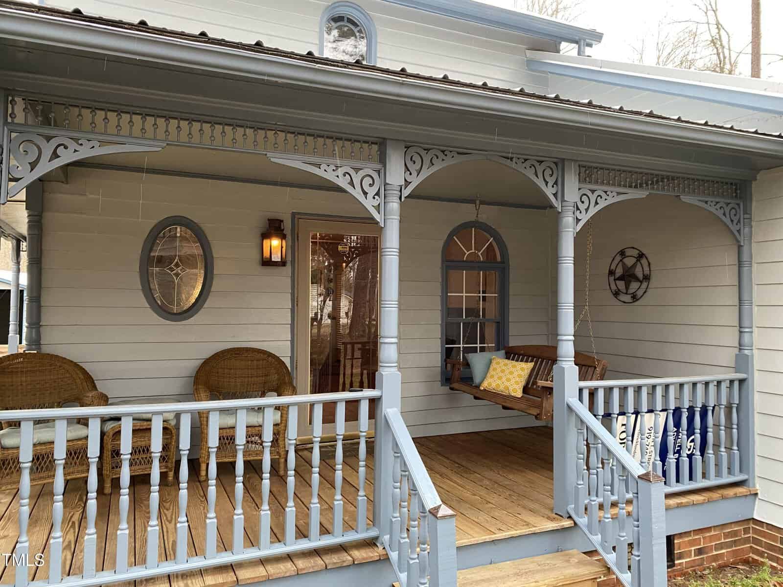 A cozy front porch with decorative white railings and intricate lattice work. the porch features wicker chairs, a wooden swing, and a round mirror on the wall of a light blue house.