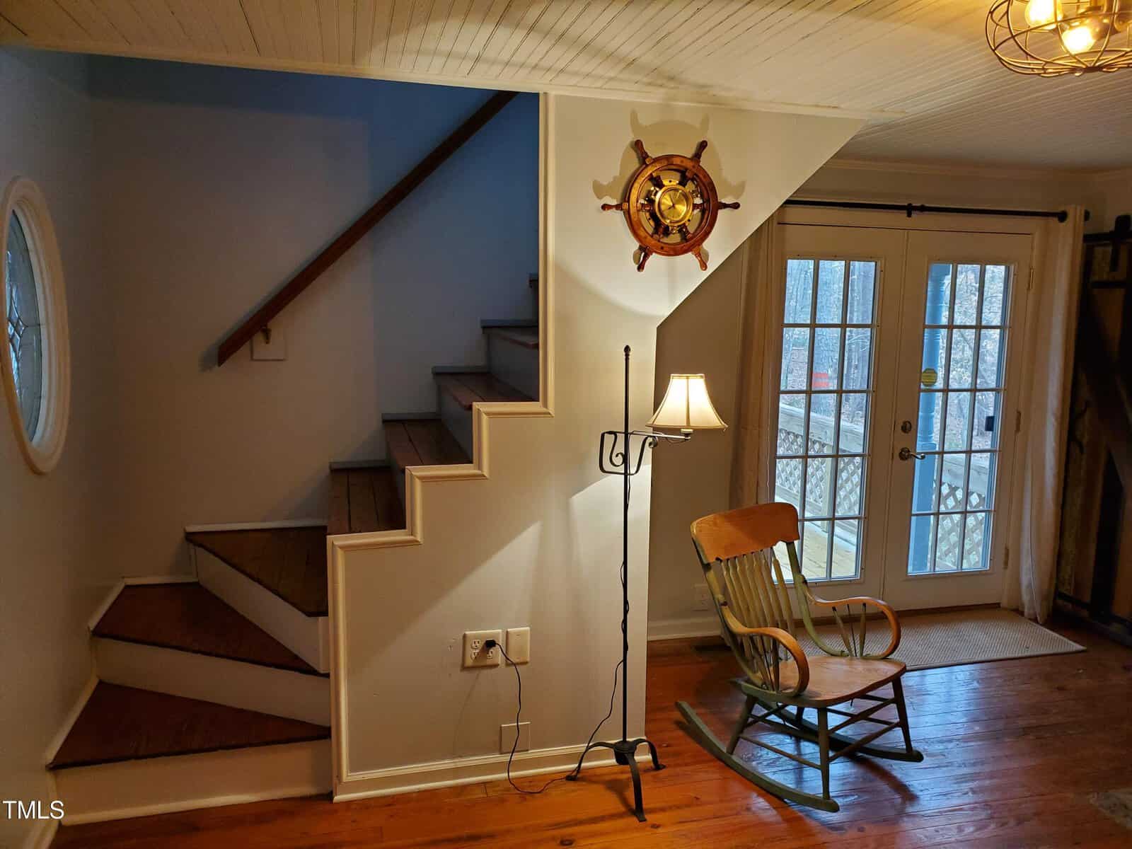 A cozy, warmly lit room featuring a wooden staircase on the left, a rocking chair, and a floor lamp. a decorative ship's wheel hangs on the wall, and a door with glass panes opens to the outside.