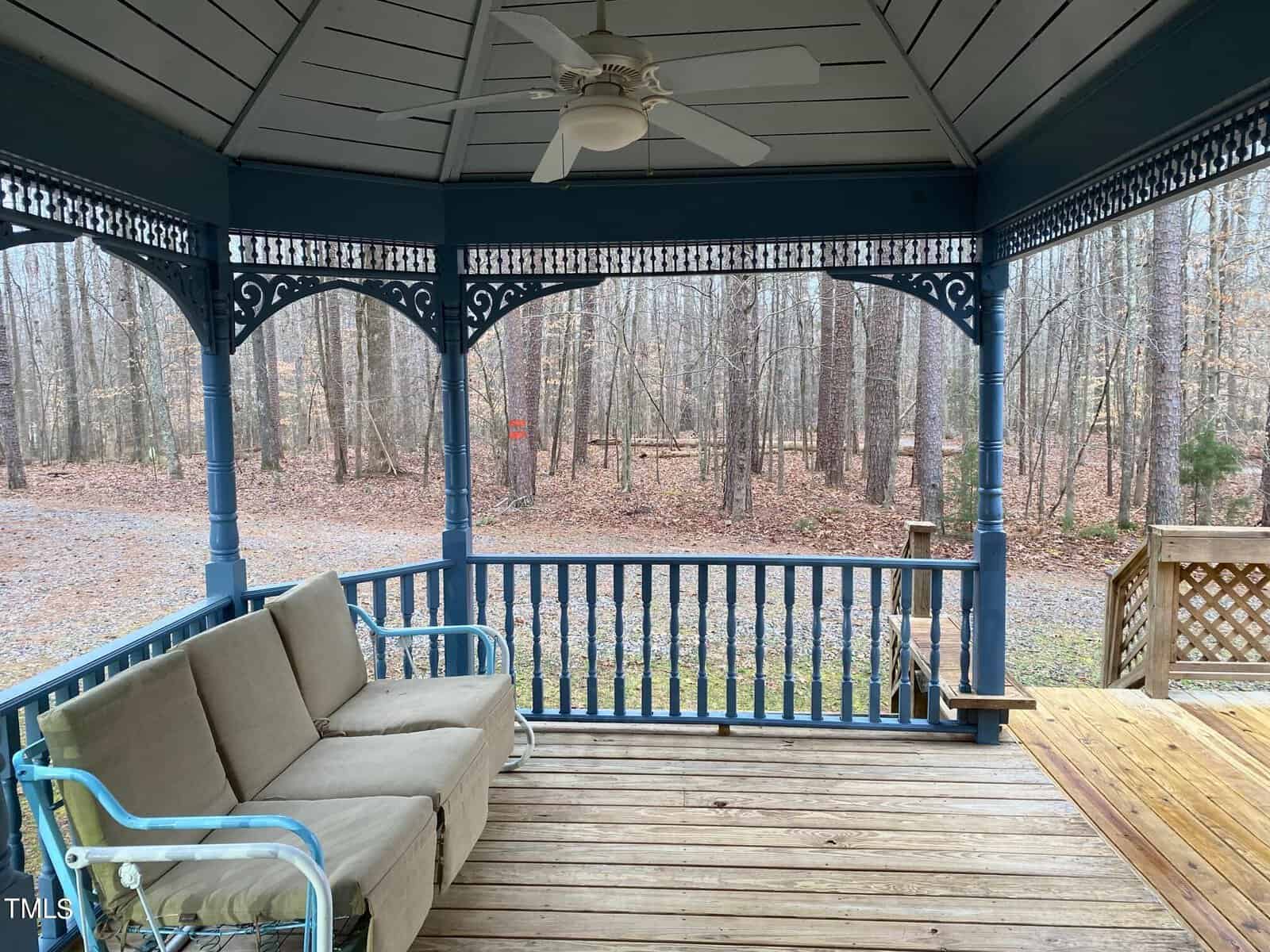 A covered wooden porch with intricate blue railings, featuring a beige sofa and a blue metal chair. the porch overlooks a leaf-covered yard with bare trees. a ceiling fan hangs above.