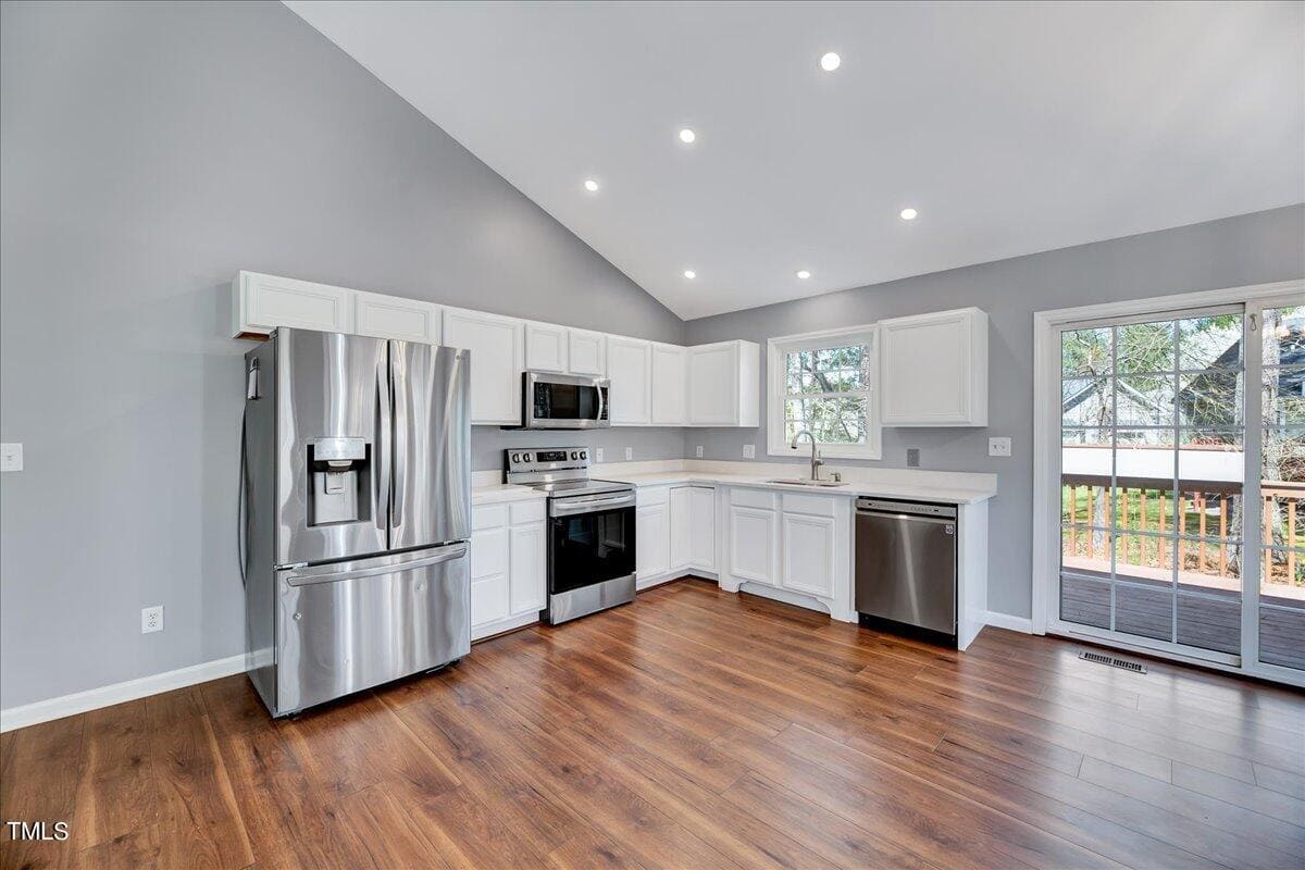 Modern kitchen with stainless steel appliances, white cabinets, wooden floors, and gray walls. Includes a refrigerator, oven, microwave, and dishwasher. A sliding glass door leads to a deck. Bright recessed lighting illuminates the space.