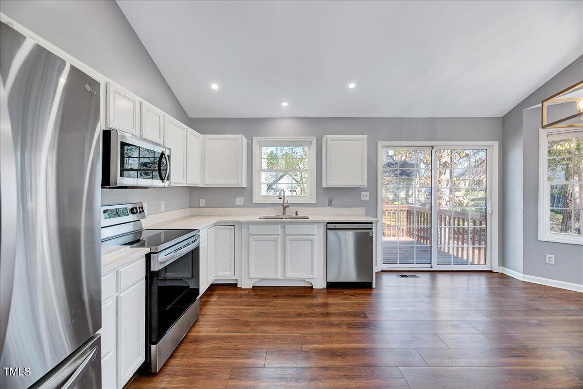 A modern kitchen with stainless steel appliances, including a refrigerator, stove, oven, and dishwasher. White cabinets and a double sink are positioned beneath large windows. The room features wooden flooring and sliding glass doors leading to a deck.
