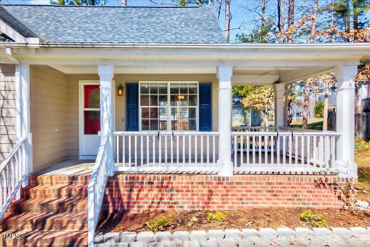 A cozy house with a front porch featuring white railings and a brick base. The porch has a red door with a window, and there's large window with blue shutters on the house. Trees and shrubs are visible in the background.