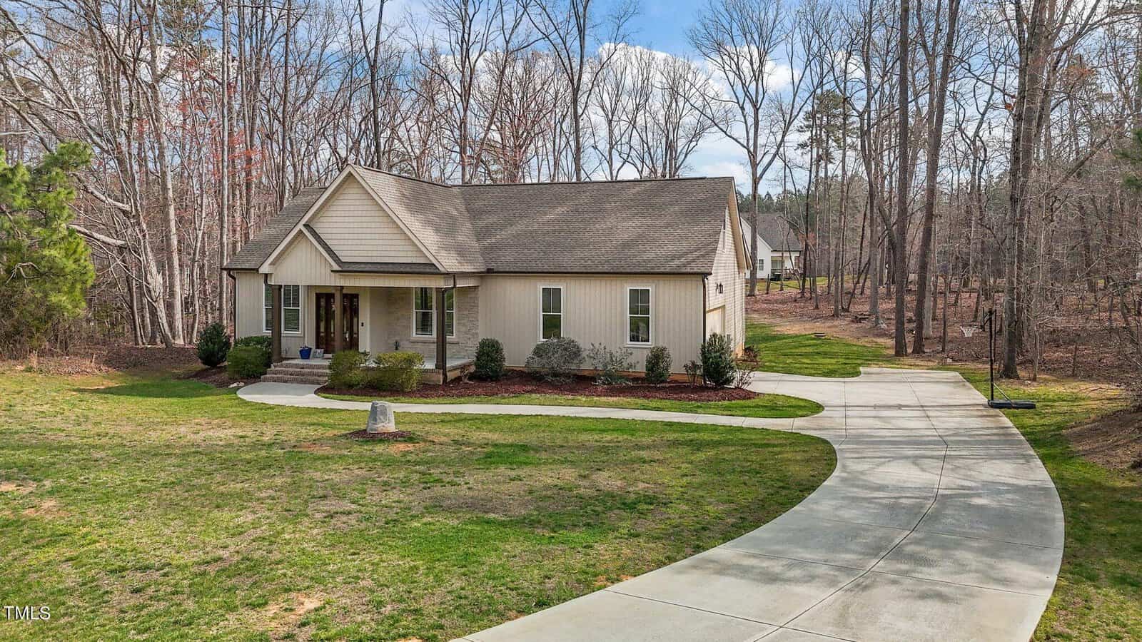 Nestled in the woods at 4334 N Greg Allen Way, Oxford, this single-story house boasts a gabled roof. A curved driveway leads to a two-car garage, surrounded by a well-maintained lawn and mature trees. The sky above is partly cloudy.