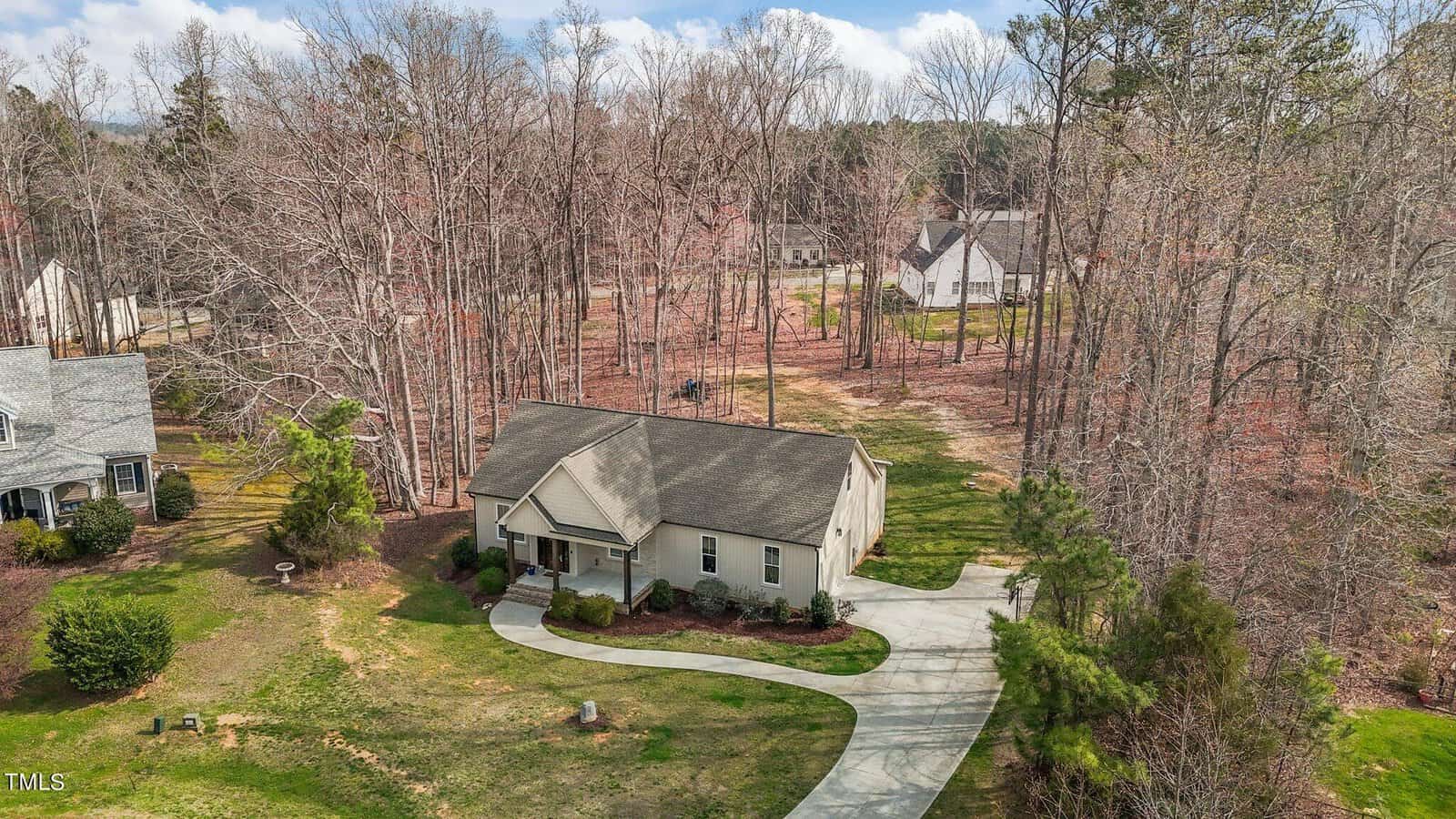 Aerial view of a single-story house with a gray roof, nestled in a wooded area. A curved driveway leads to the front porch. Surrounding the house are bare trees, a manicured lawn, and neighboring homes in the background.