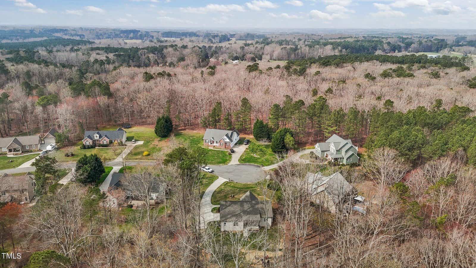Aerial view of a residential neighborhood surrounded by dense forest. Several houses are situated on a cul-de-sac, with trees shedding leaves, indicating a late autumn setting. The landscape extends into rolling hills under a cloudy sky.