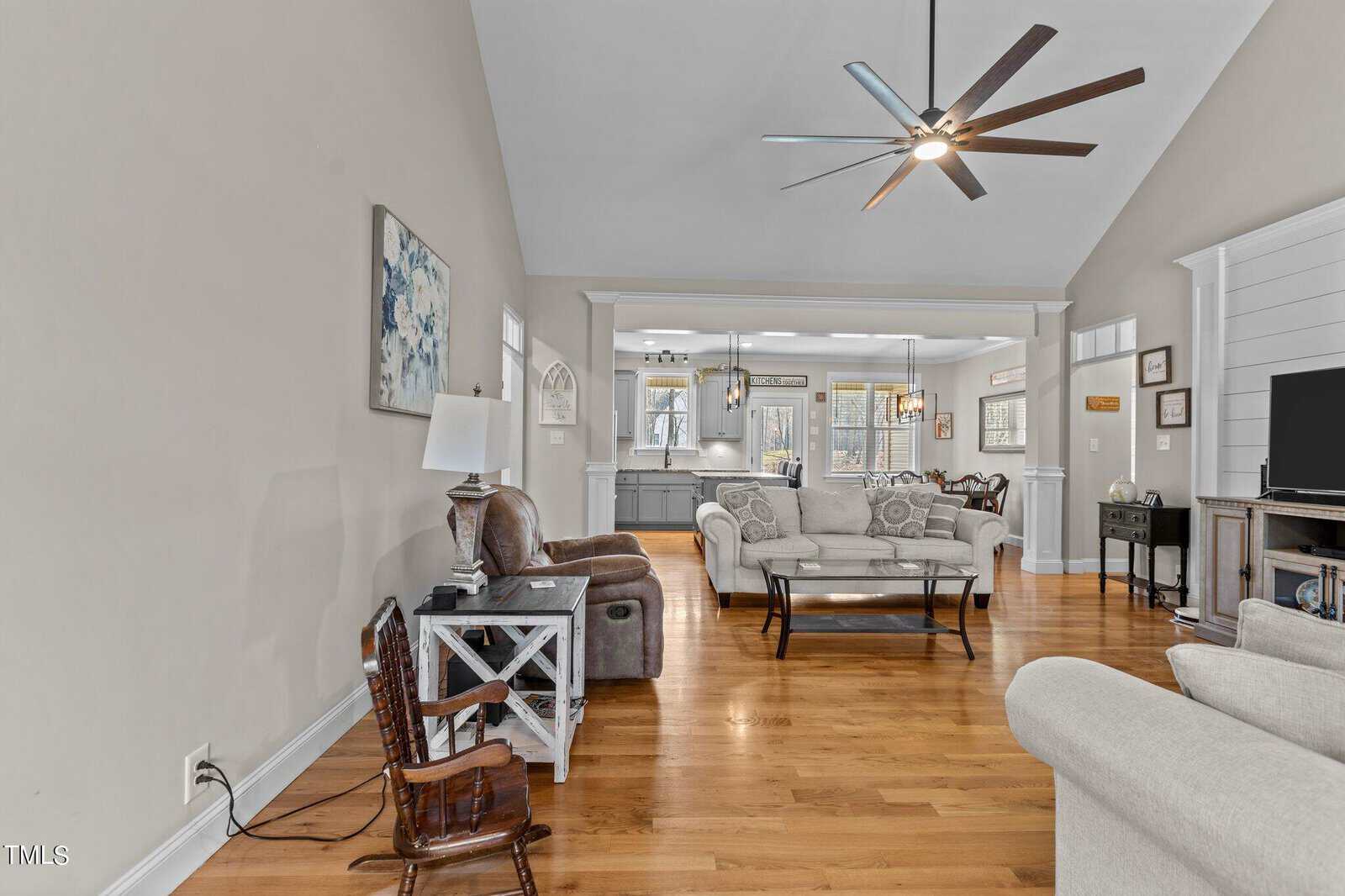 Spacious, modern living room with vaulted ceiling, light beige walls, and wooden floors. It features a large fan, a mix of armchairs and sofas, and a seamless transition to a bright kitchen area in the background.
