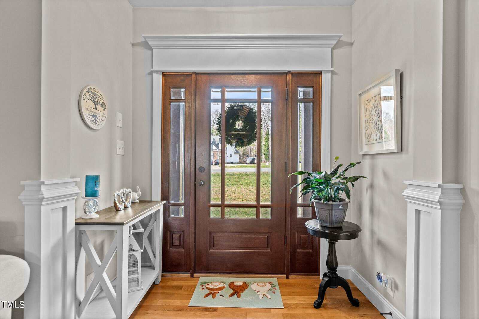 A welcoming entryway with a wooden front door featuring glass panels. A small table on the left holds decorative items, and a potted plant is on the right. A colorful doormat with animal designs lies on the wooden floor.