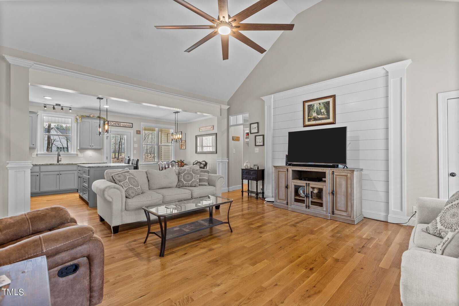 A modern living room with hardwood floors, a ceiling fan, and recessed lighting. It features a flat-screen TV on a wooden stand, a beige sofa, a brown recliner, and glass-top coffee tables. The open kitchen is visible in the background.