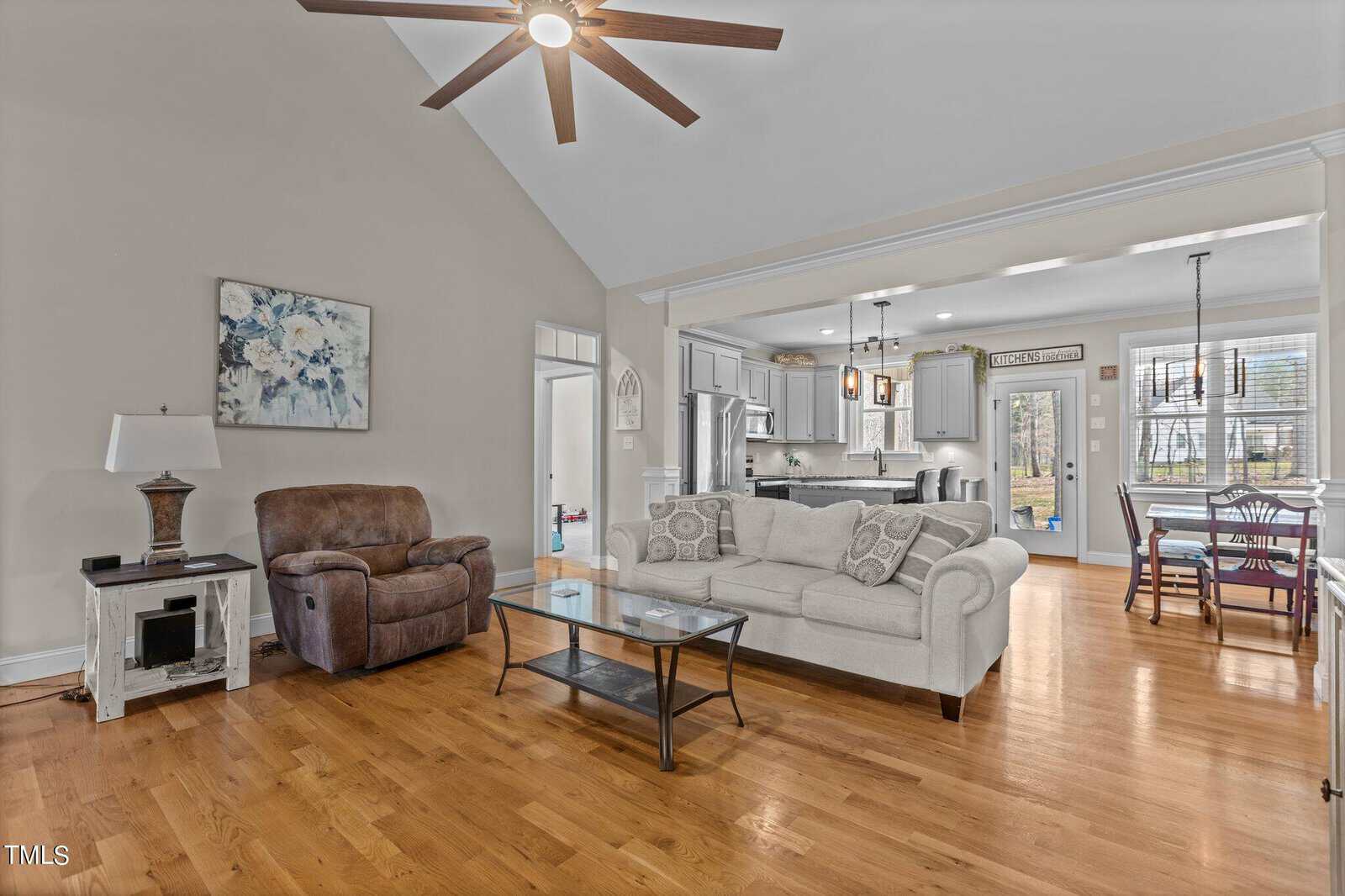 A spacious living room with wooden floors, featuring a beige sofa, glass coffee table, and a brown recliner. There's a modern kitchen in the background with light gray cabinets and a dining area adjacent. A ceiling fan and decorative wall art are visible.