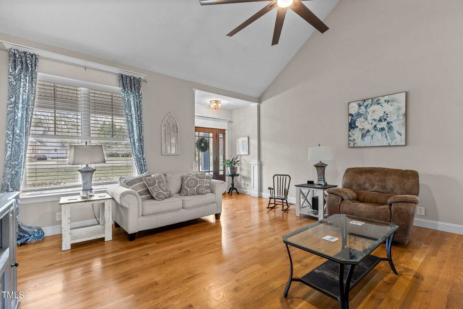 A cozy living room with a vaulted ceiling, featuring a beige sofa, brown armchair, and glass coffee table on a polished wooden floor. Blue curtains and a floral painting add color, while large windows allow daylight to fill the space.