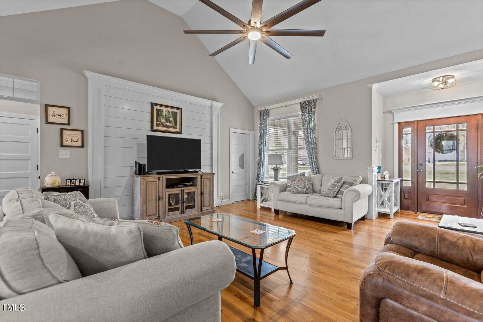 A cozy living room with vaulted ceilings, featuring a large ceiling fan, two beige sofas, a brown recliner, and a glass coffee table. A TV sits on the cabinet, and large windows let in natural light. The entryway has a wooden door and side windows.
