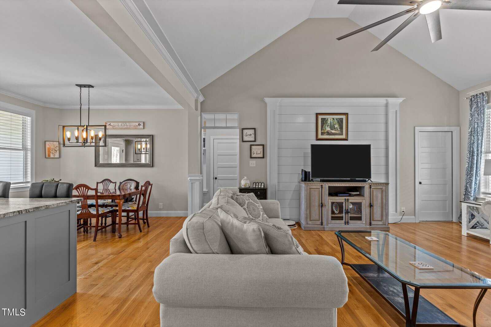 A spacious living area with a vaulted ceiling, featuring a gray sectional sofa, a glass coffee table, and a TV on a wooden stand. A dining area with a wooden table and chairs is in the background, and decorative lighting hangs above.