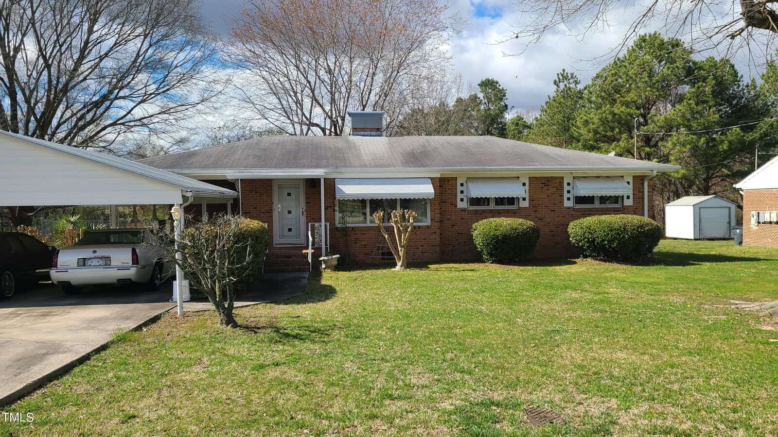 A single-story brick house with a white carport on the left. there's a well-kept lawn in front and a white detached garage to the right under a clear blue sky.