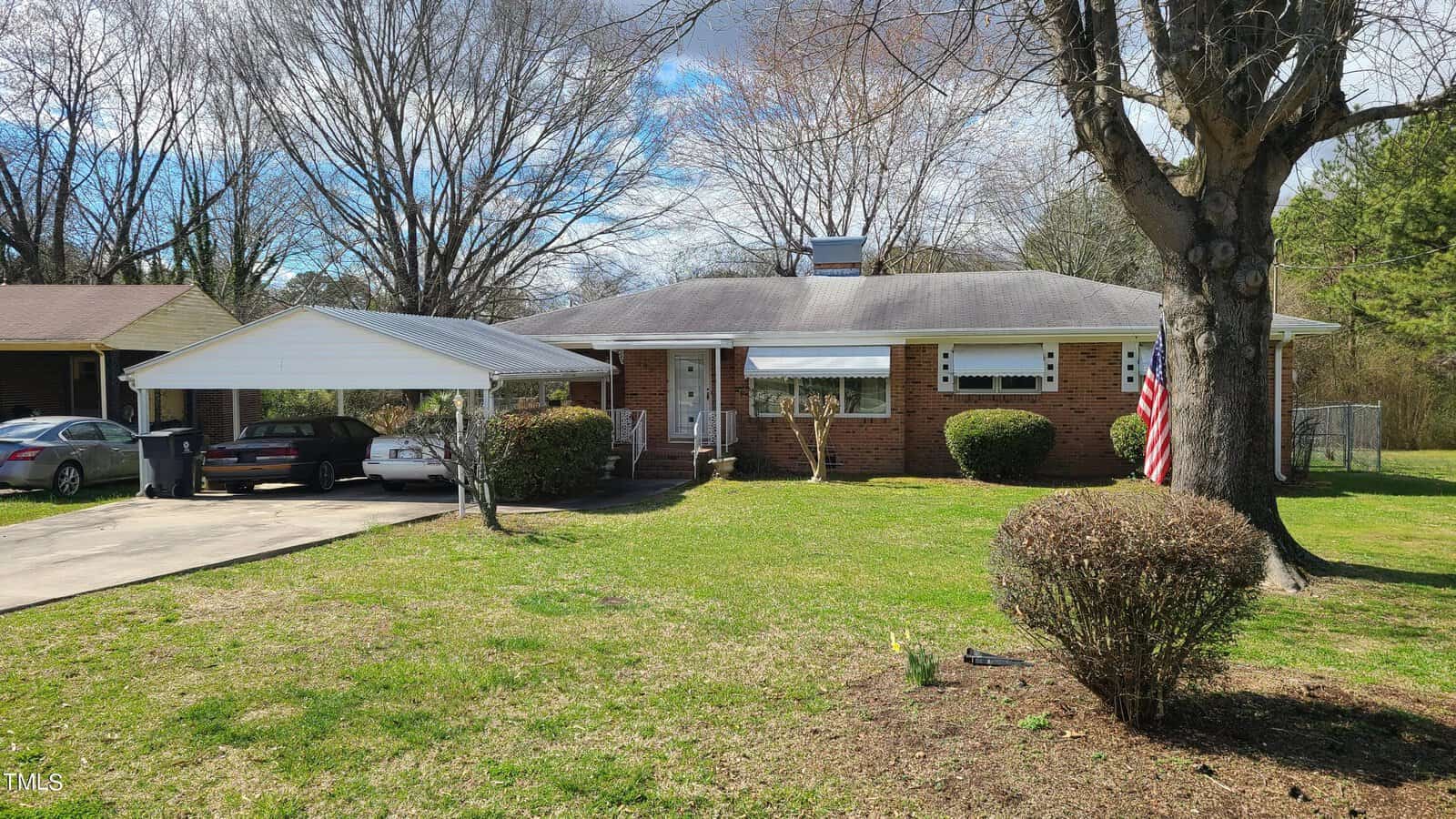 A one-story brick house with a carport on the left, featuring a well-kept lawn and an american flag hanging on the right. there's a large tree to the right and a sedan parked under the carport.