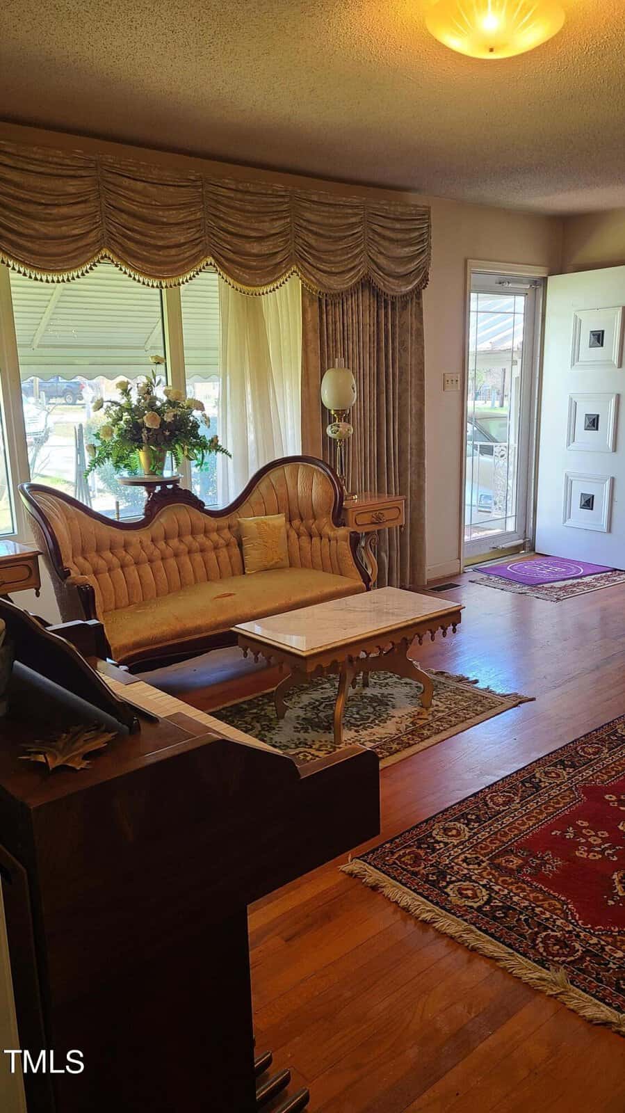Interior of a living room with a vintage-style beige sofa, wooden coffee table, oriental rugs, and a window with sheer curtains and ornate valances. bright natural light illuminates the space.
