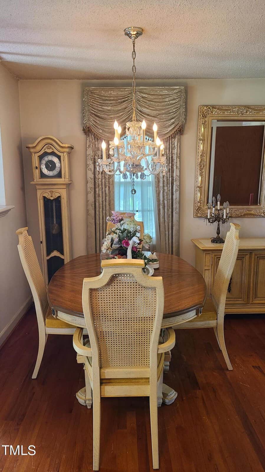 A traditional dining room featuring a round wooden table with elegant white chairs, an ornate chandelier above, and surrounded by golden-toned furniture including a mirror and a cabinet.