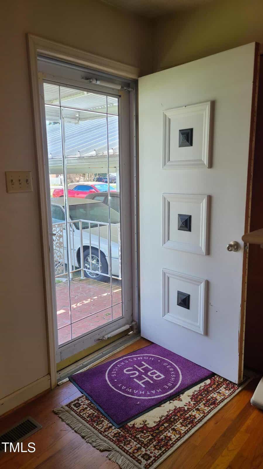 A residential entryway featuring a white door with built-in blinds, surrounded by a white frame and flanked by light-colored walls. a decorative rug rests on the wooden floor in front of the door.
