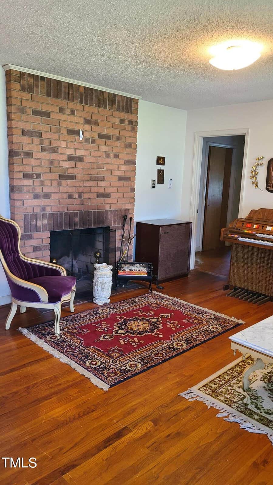 A cozy living room featuring a brown brick fireplace, a patterned red rug, and a purple armchair. hardwood floors and subtle lighting create a welcoming space.