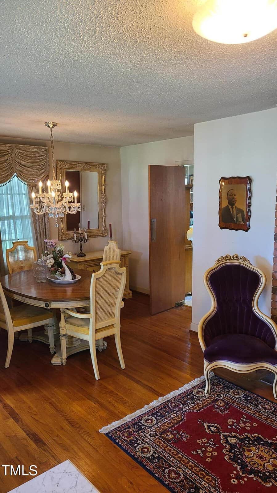 Interior of a home showing a dining area with a wooden table, white chairs, and a chandelier. an ornate purple chair and a portrait hanging on a wall are also visible. a persian rug is on the floor.