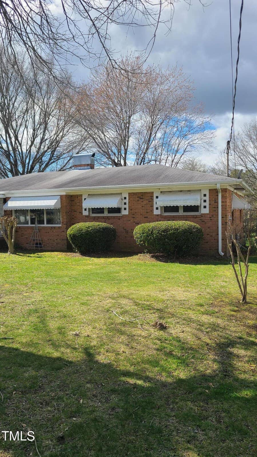 A single-story brick house with white trim and three front windows, partially obscured by two large, manicured bushes. the house features a white awning and a lawn with scattered leaves.