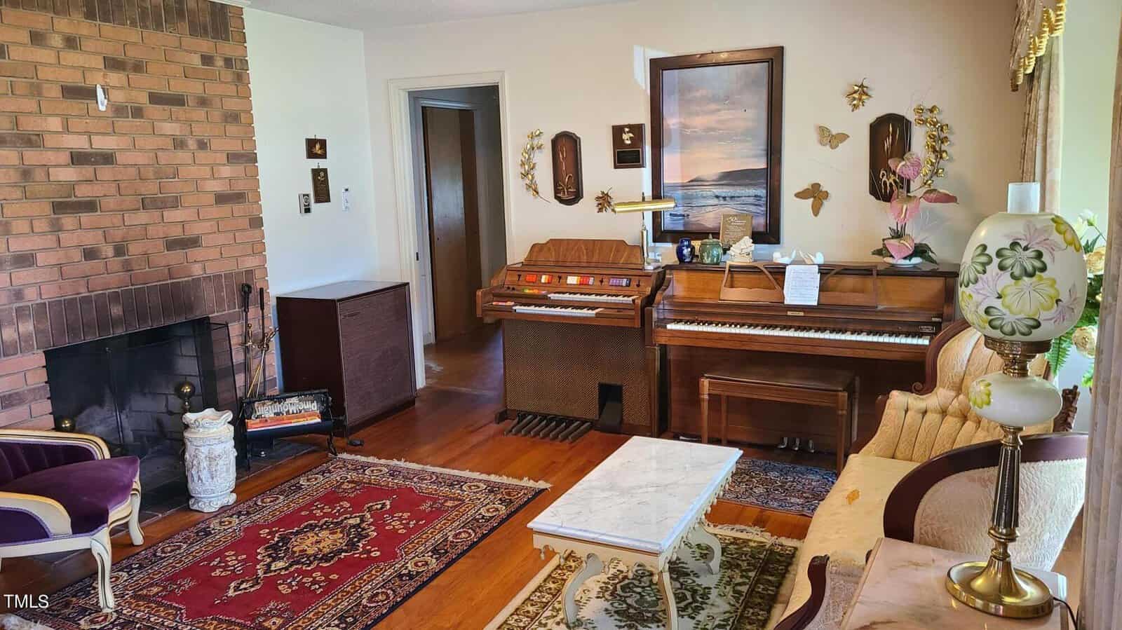 Interior of a cozy living room featuring a brick fireplace, an upright piano, decorative lamps, a printed rug, and various wall ornaments including paintings and metal sculptures.