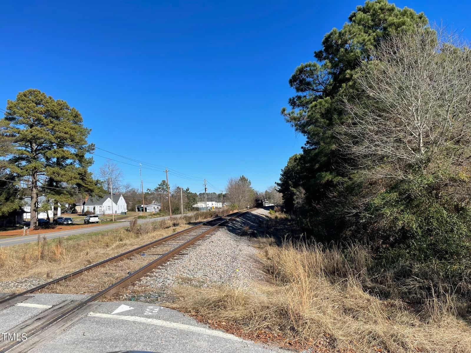 Railroad tracks cutting through dry grass, flanked by evergreen trees and a road with cars and houses under a clear blue sky.