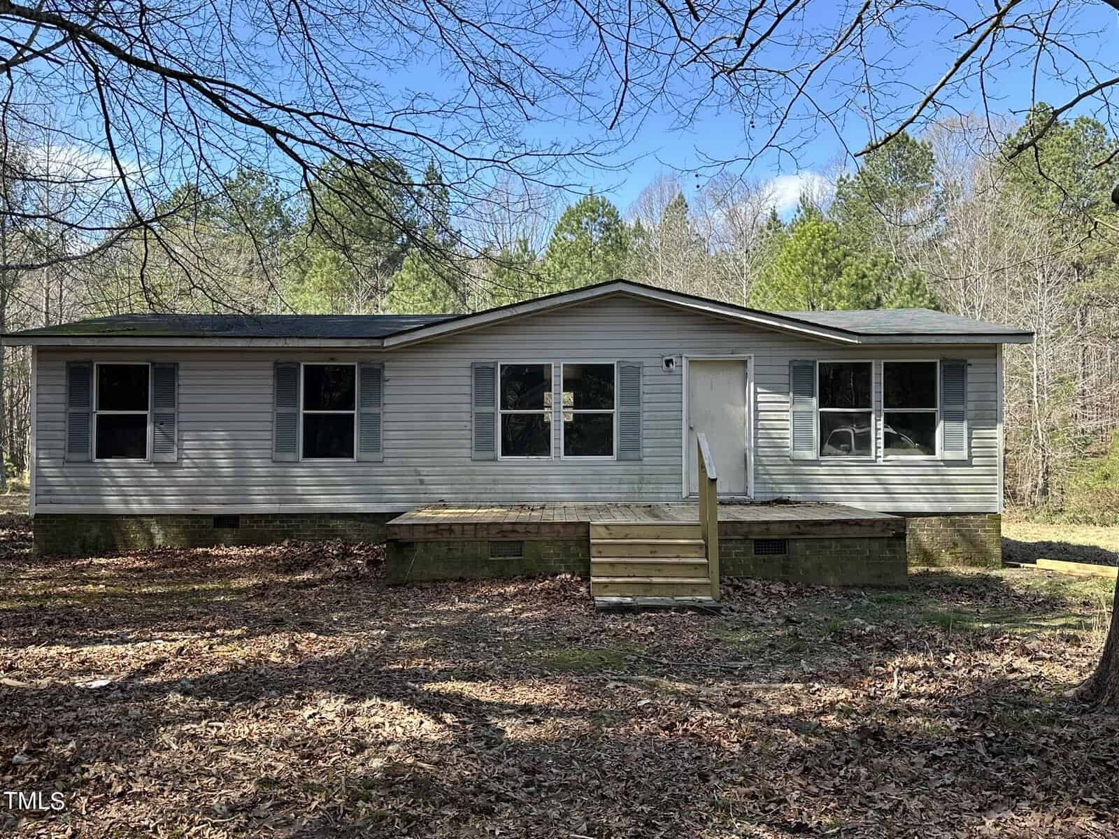 An off-white mobile home at 4131 Doe Court features a small wooden front porch set amidst the wooded surroundings of Oxford. The ground is covered with dry leaves, and trees with bare branches encircle the home, while the sky remains partly cloudy.
