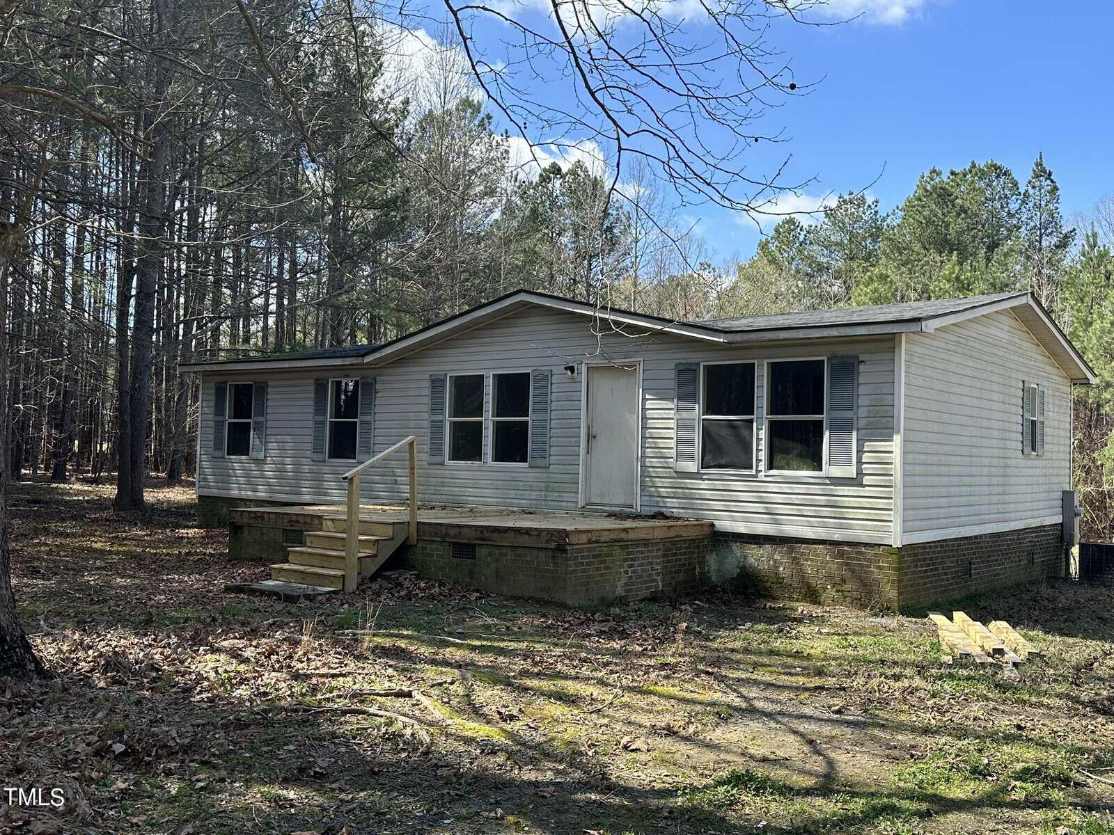 A single-story house with light-colored siding and a front porch with steps. It is surrounded by tall trees and set in a grassy area under a partly cloudy sky. A few wooden planks are lying on the ground near the porch.