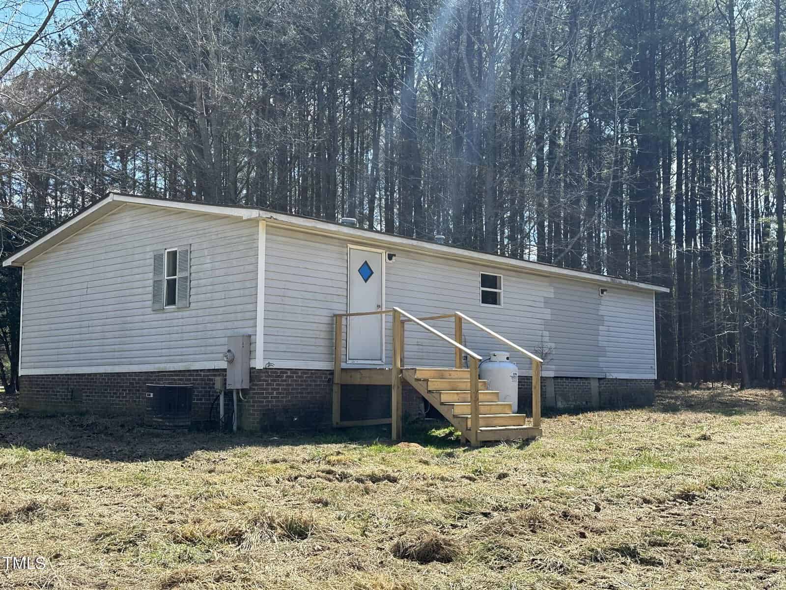 A small white mobile home with a pitched roof sits elevated on a foundation of brick and wooden steps. It is surrounded by a grassy area with tall trees in the background under a clear sky.