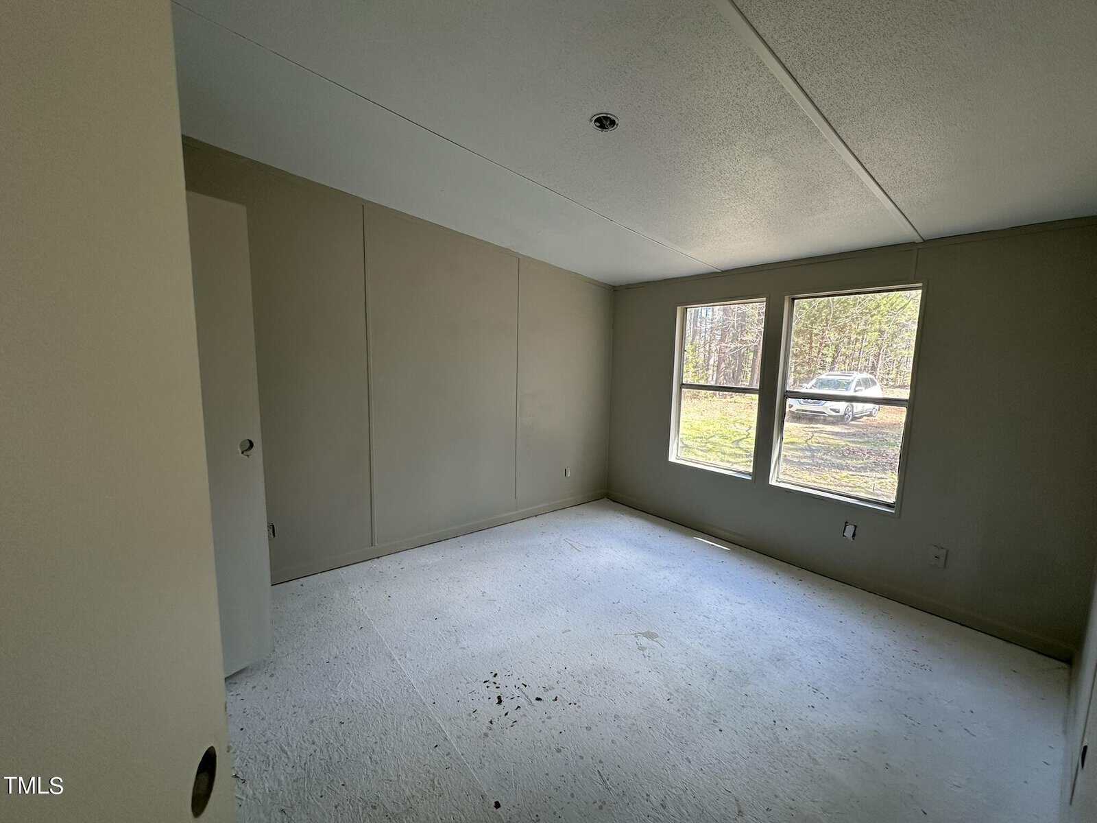An empty room with light-colored walls, a door slightly ajar on the left, and two large windows on the right letting in natural light. Outside, a car is visible with trees in the background. The floor is bare and unfinished.