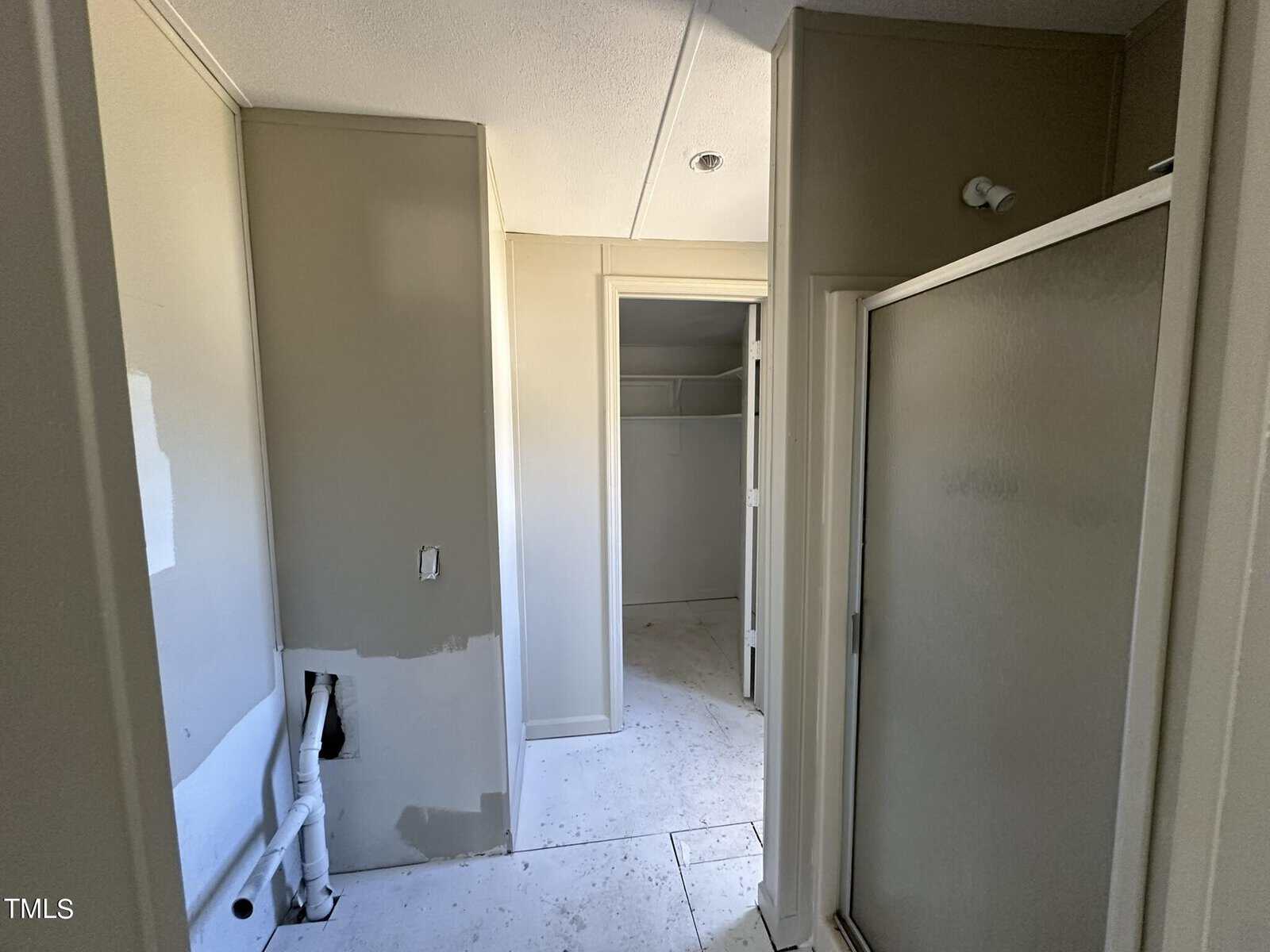 A partially completed bathroom with unpainted walls, an open closet door in the background, and an unfinished plumbing area on the left. A shower stall with a frosted door is on the right. The floor is bare with visible patches.
