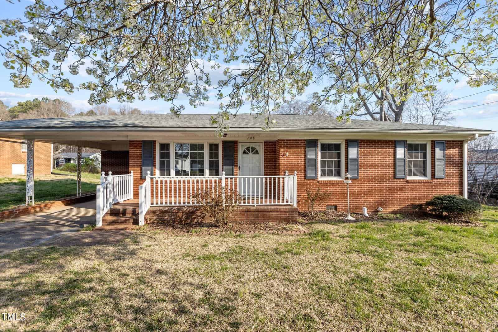 Located at 208 Grove Street in Oxford, this single-story brick house boasts white shutters and a charming front porch beneath a large blooming tree. The porch, with its railing and steps, leads to a walkway. A driveway on the left extends under a carport attached to the home, all surrounded by an expansive grassy yard.