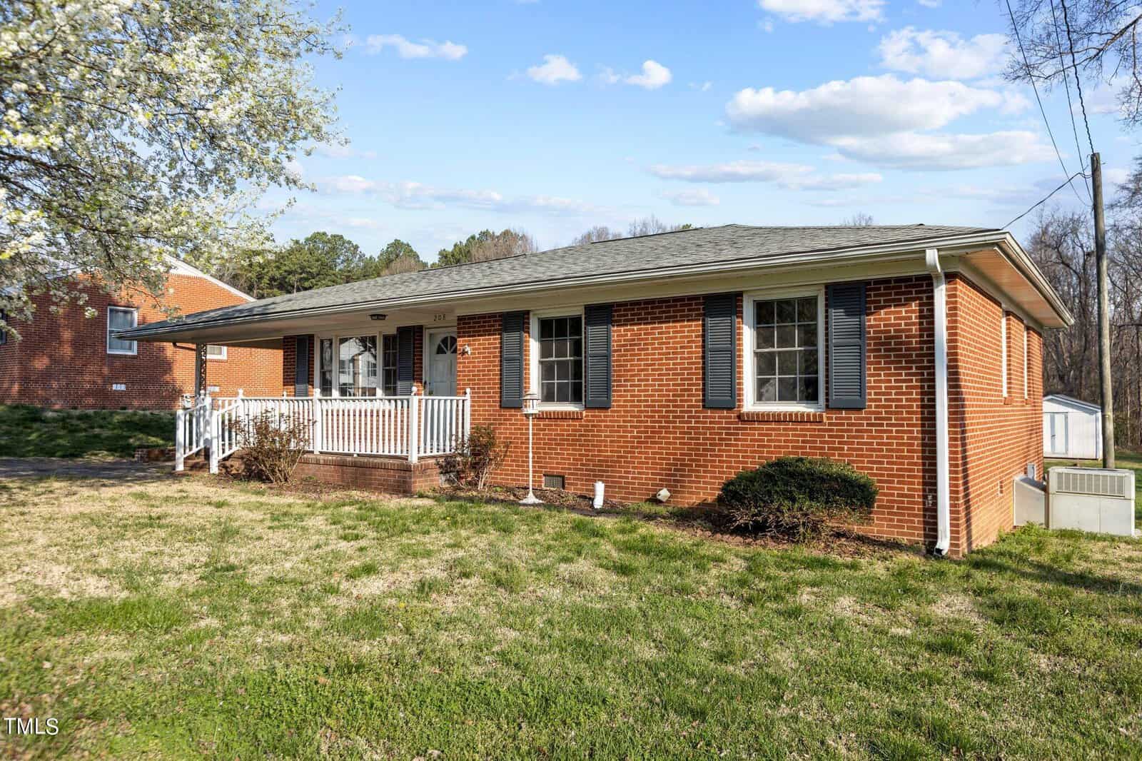 A single-story red brick house with a covered front porch and white columns. The yard is neatly maintained with green grass and a few bushes. A flowering tree is in the foreground, and the sky is partly cloudy with some blue patches.