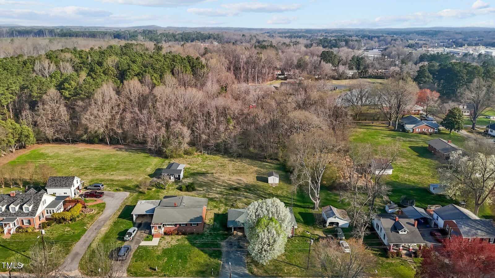Aerial view of a suburban neighborhood with houses surrounded by trees and grassy areas. The landscape includes a mix of dense woods and open lawns, with a clear blue sky above.