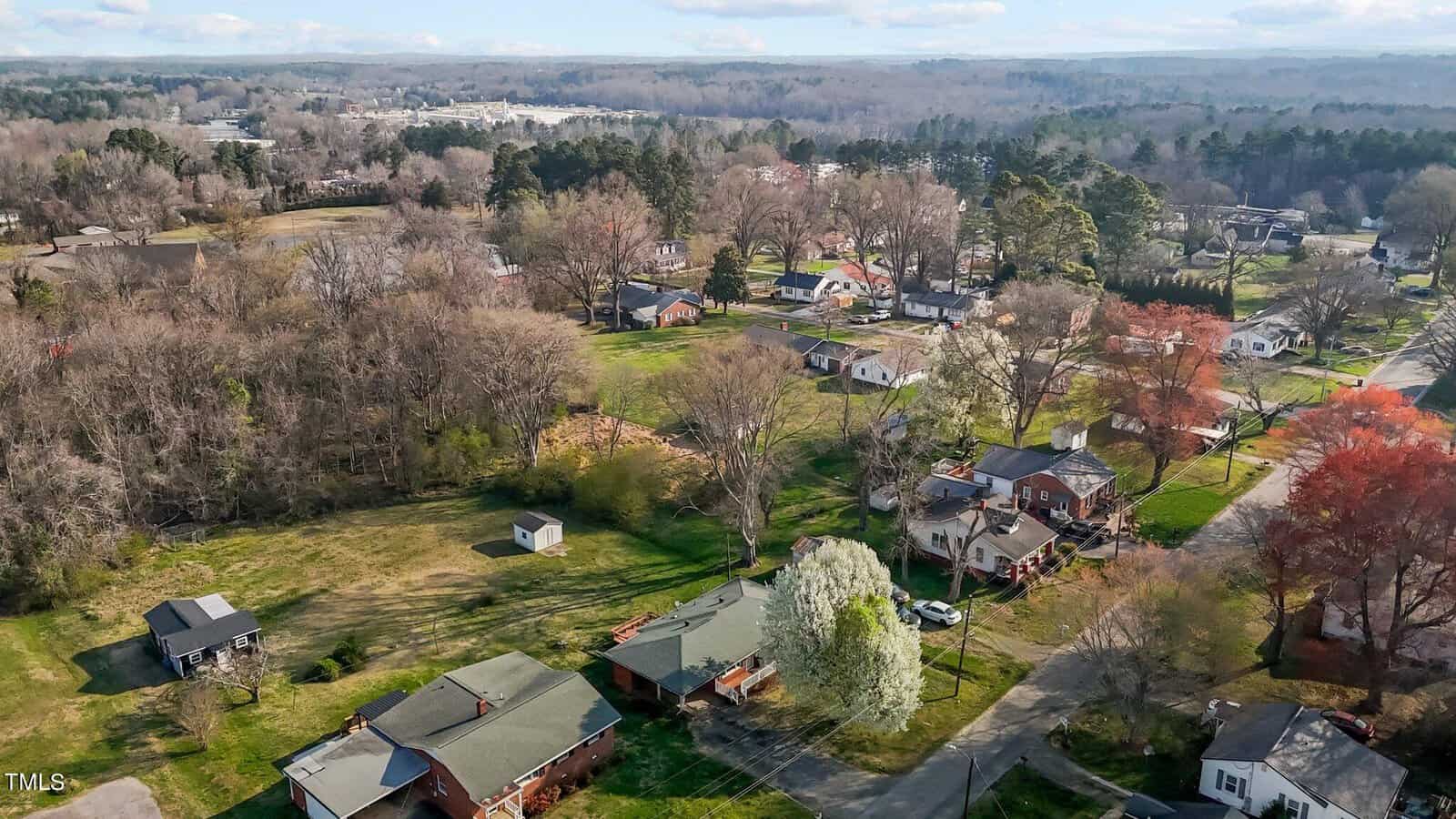 Aerial view of a suburban neighborhood featuring a mix of houses, trees with autumn hues, and open green spaces. The landscape is dotted with roads, a few small structures, and surrounded by dense trees in the distance under a partly cloudy sky.