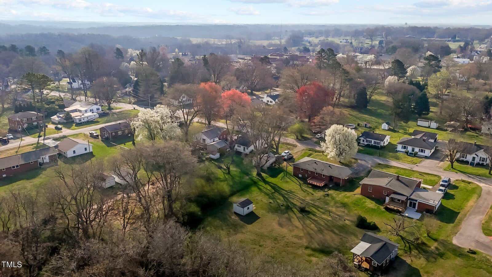 Aerial view of a suburban neighborhood in early spring, featuring houses with yards, trees with red and white blossoms, and a distant view of more trees and fields under a blue sky with scattered clouds.