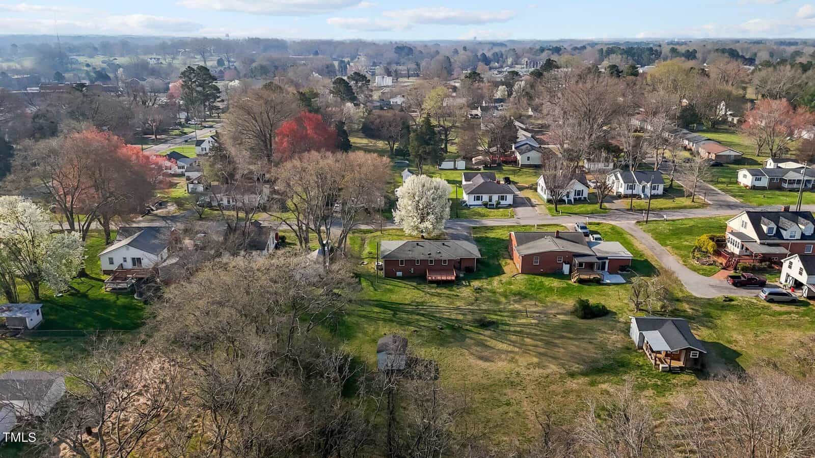 Aerial view of a residential neighborhood with houses surrounded by trees. The landscape features a mix of bare and blooming trees, indicating early spring. Roads wind through the area, and more greenery can be seen in the distance.