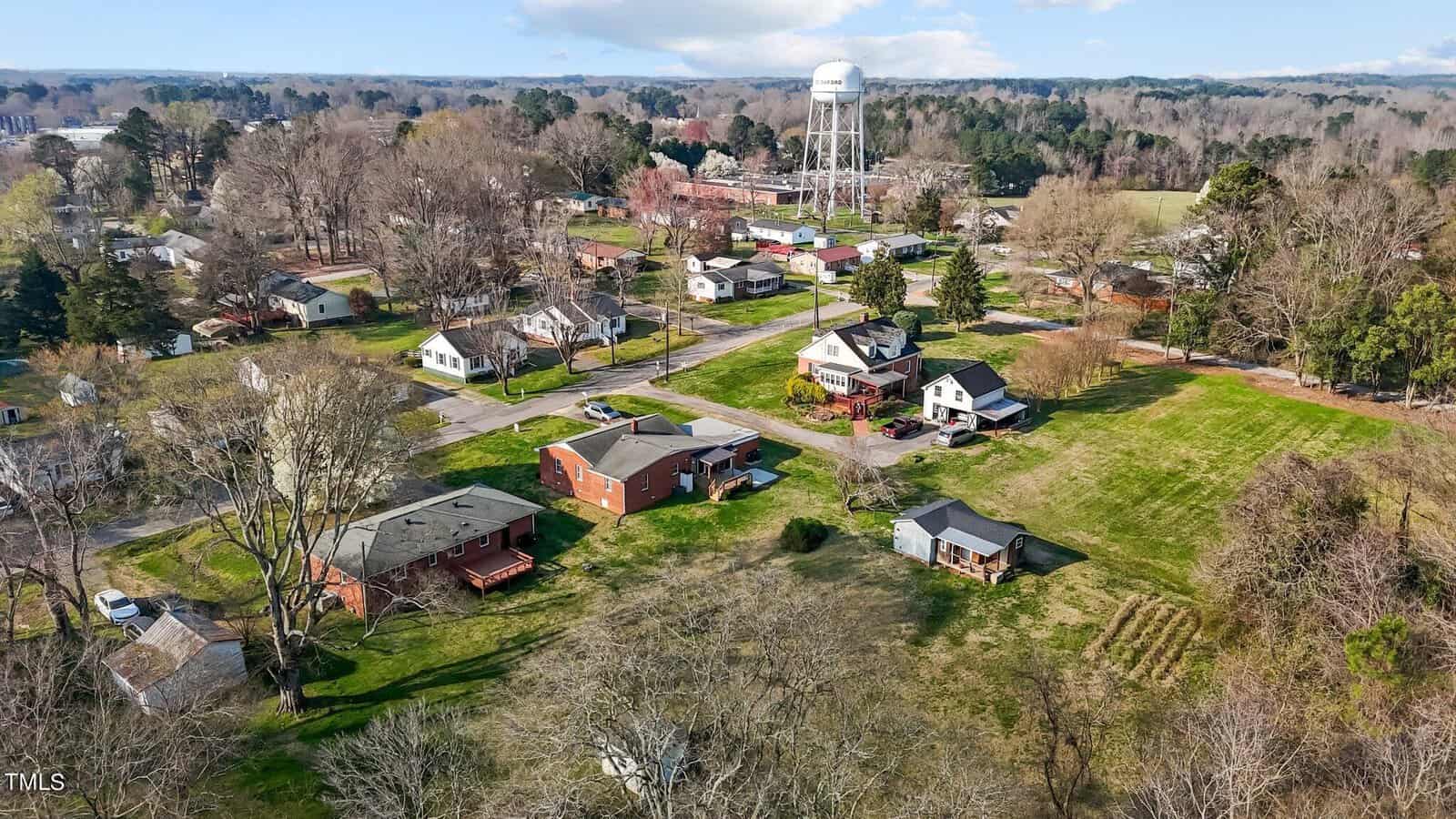 Aerial view of a suburban neighborhood with a mix of houses, green lawns, and trees. In the background, there is a large water tower and more residential homes surrounded by wooded areas under a slightly cloudy sky.