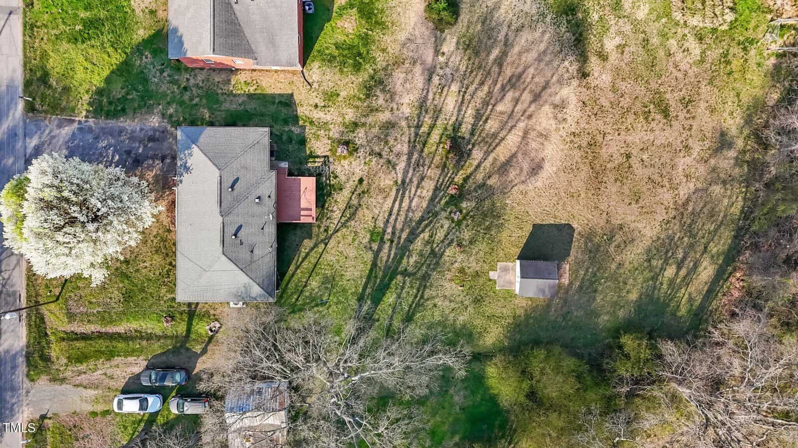 Aerial view of a residential area with a beige house and a small shed in a backyard. Three cars are parked near the house. The yard is mostly grass with some trees casting long shadows on the ground.